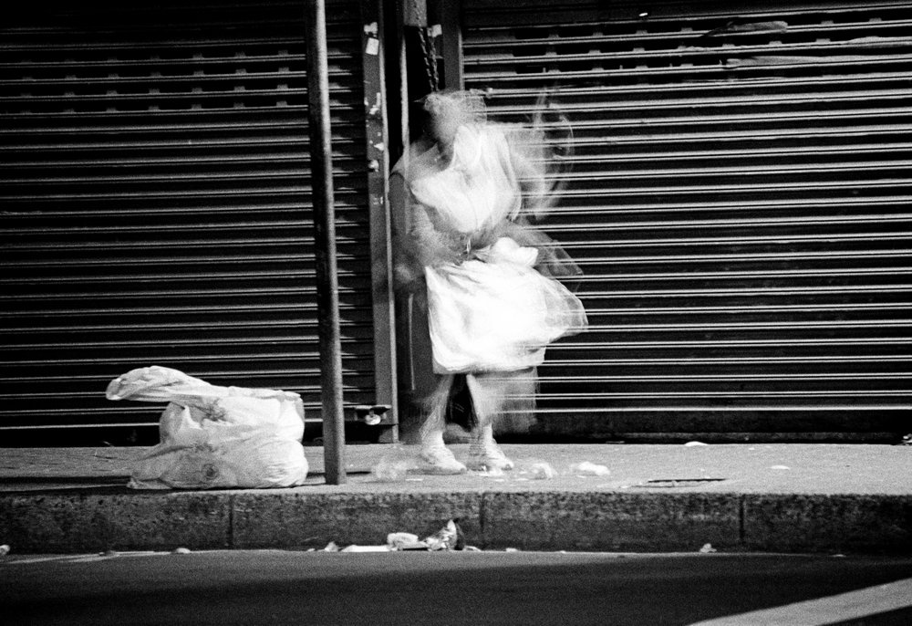 Sabelo Mlangeni's monochrome photographic print 'Invisible Woman I' shows a woman walking on a concrete sidewalk.
