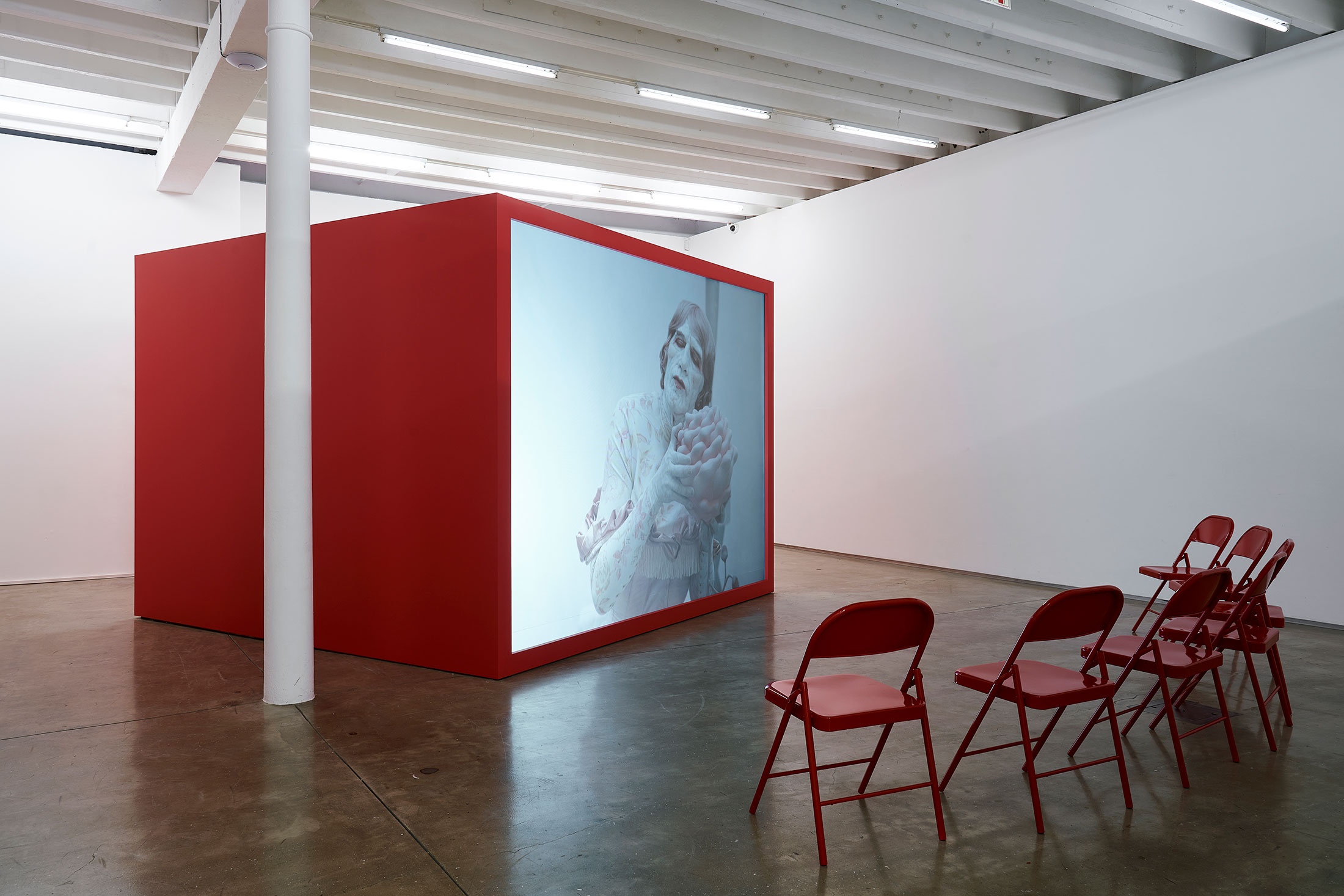Installation photograph from the 'A Little After This' exhibition in A4 Arts Foundation's gallery that shows Alex Da Corte's video installation 'ROY G BIV'. On the left, a large red wooden box with a back-projected screen plays Da Corte's video. On the right, 7 red powder-coated viewing chairs are arranged in an arch.
