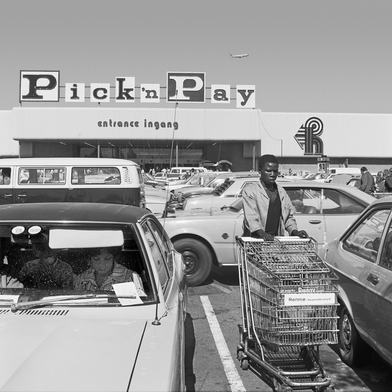 David Goldblatt's monochrome photograph 'Hypermarket employee collecting trolleys, Boksburg' shows an individual pushing a trolley in a parking lot.
