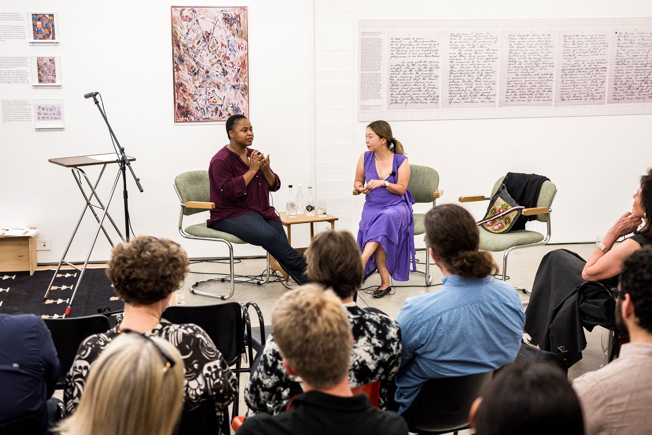 Event photograph from the Ernest Mancoba Symposium at A4 Arts Foundation. At the front, audience members are seated in rows. In the middle, two speakers are seated facing the audience. At the back, posters of artworks and text line a white wall.
