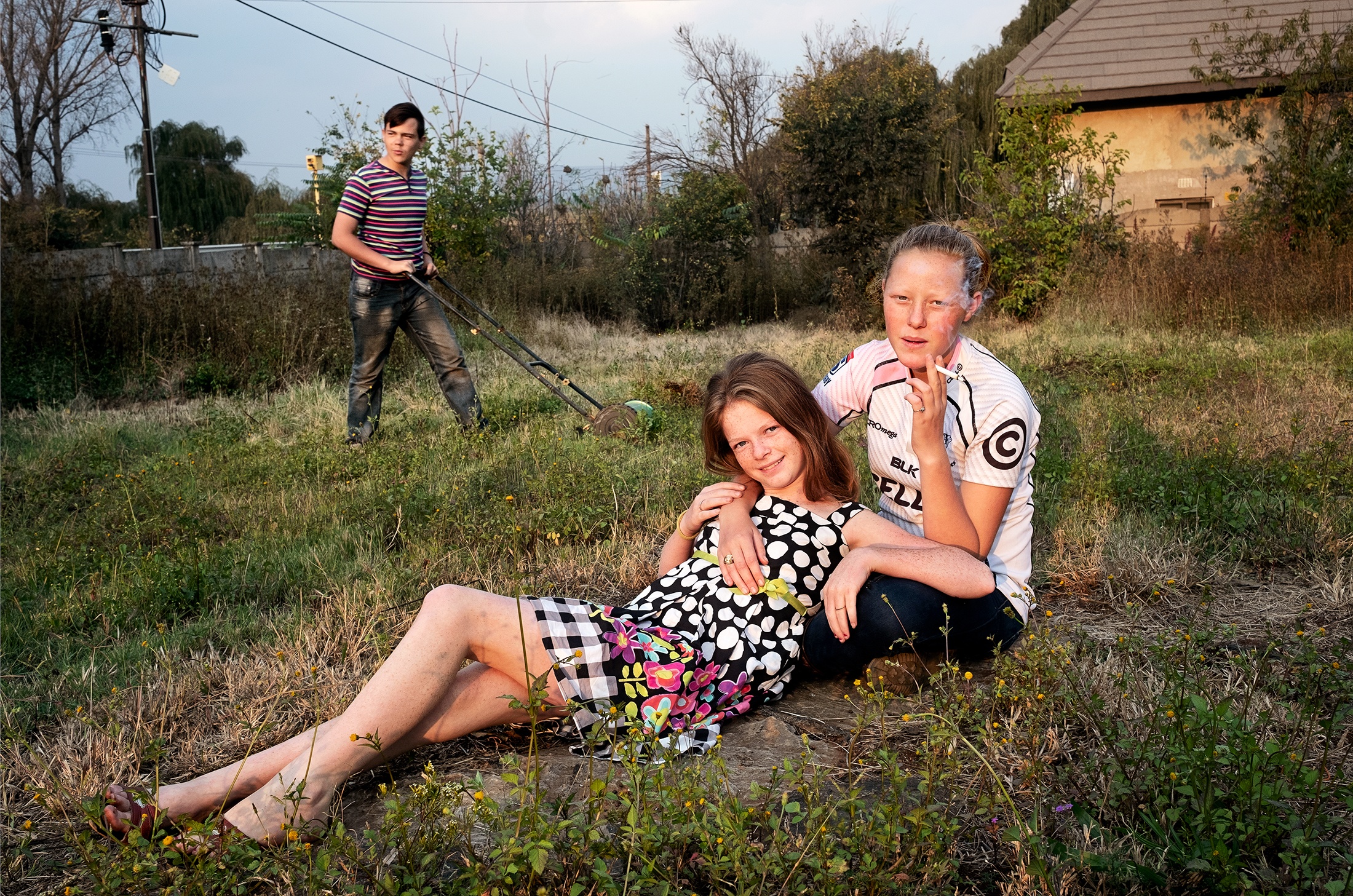 Lindokuhle Sobekwa's photograph 'Jordan and Nadine' shows two children sharing a cigarette on a stretch of grass, with another in the background operating a lawnmower.
