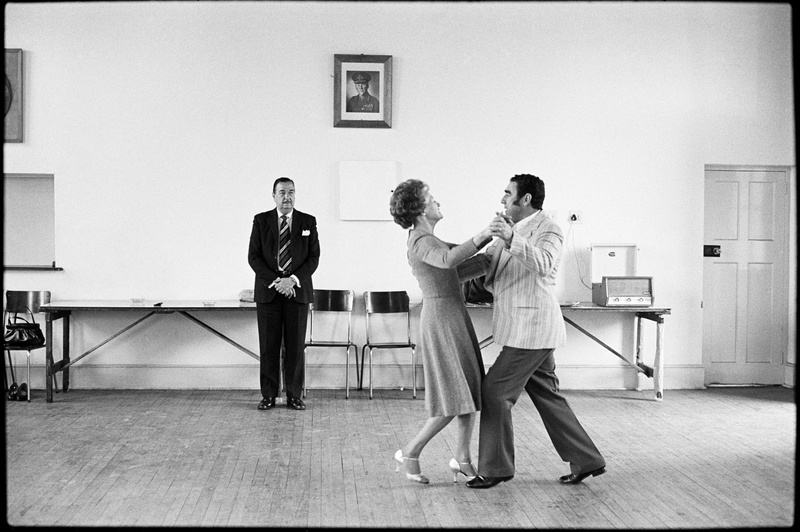 David Goldblatt's monochrome photograph 'Dancing-master Ted van Rensburg watches two of his ballroom pupils, swinging to a record of Victor Sylvester and his Orchestra, in the MOTHS’ Hall at the old Court House, Boksburg' shows two individuals dancing in a hall with a wooden floor.
