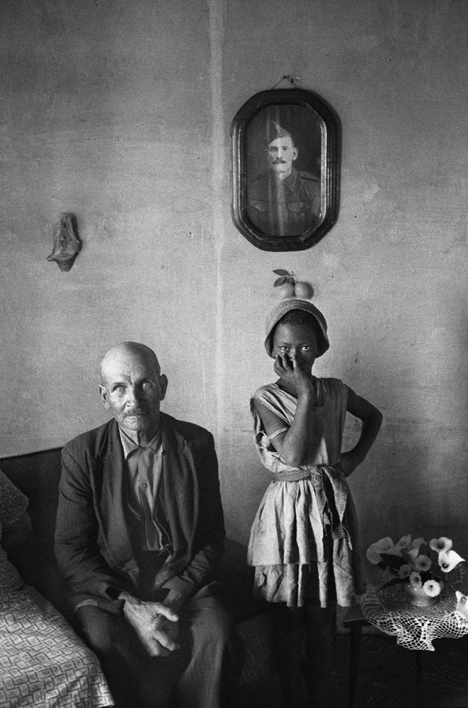David Goldblatt's black-and-white photograph 'A plot-holder with the daughter of his servant, Wheatlands, Randfontein' shows a man seated on a couch and next to him, a young girl standing.
