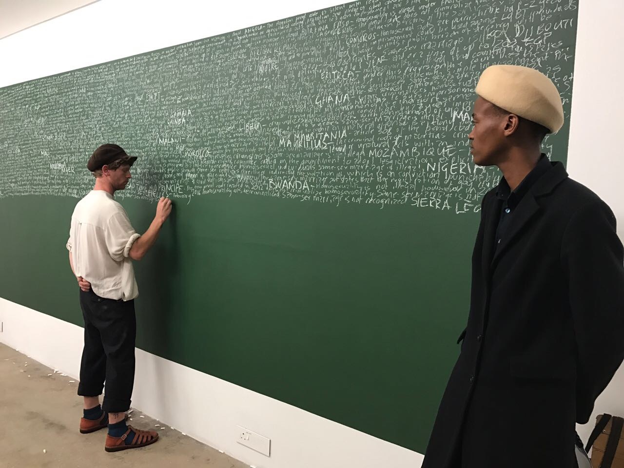 Photograph of Luvuyo Nyawose and Brett Charles Seiler's performance installation 'Reading Homophobia'. On the right, Nyawose is standing. On the left, Seiler writes on a green chalkboard.
