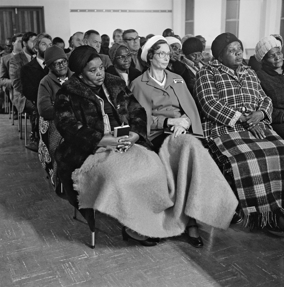 David Goldblatt's monochrome photograph 'Methodists meet to find ways of reducing the racial, cultural and class barriers that divide them' shows individuals seated on chairs in rows indoors.
