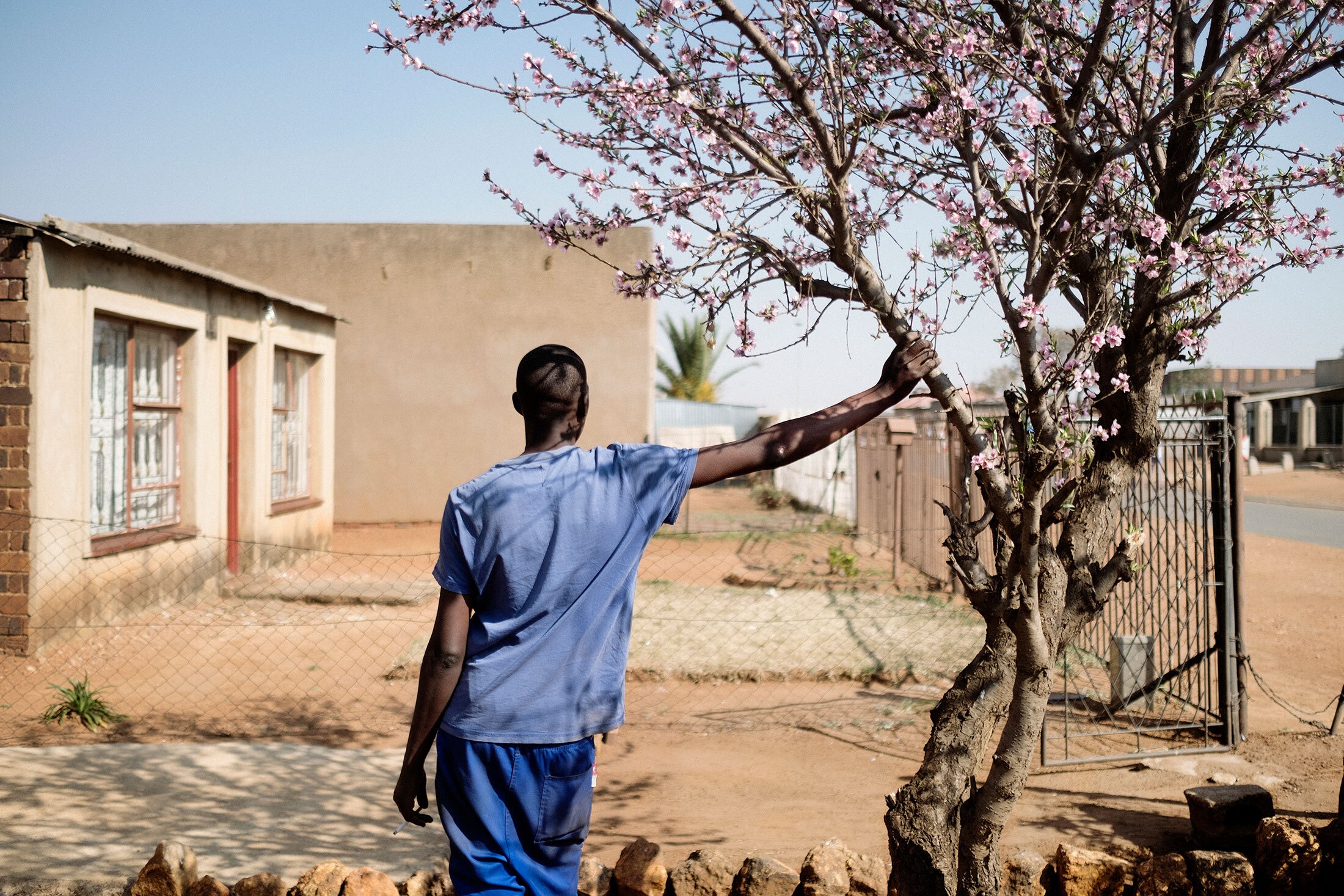 Lindokuhle Sobekwa’s photograph ‘Mzwandile on rehabilitation’ (2019) depicts a person standing in a house’s front yard steadying themselves against a tree branch.
