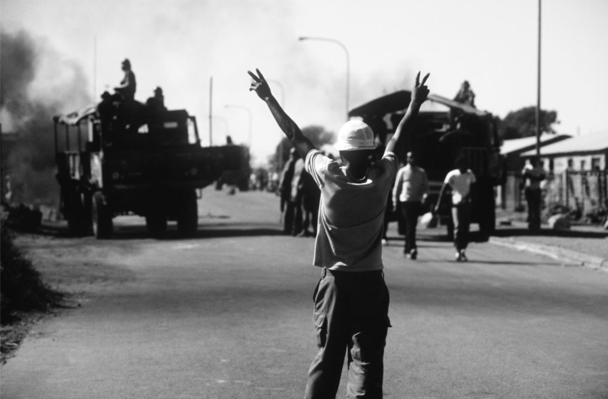 Peter Magubane’s monochrome photograph ‘Give peace a chance, Soweto’ shows an individual standing in a road making peace signs with both hands held high at the front, and large vehicles at the back.

