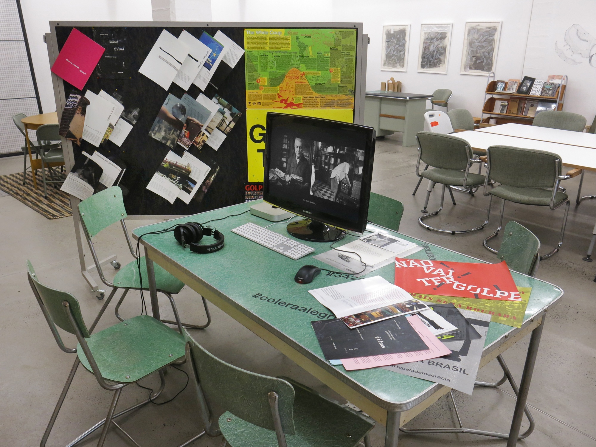 Installation photograph from the ‘How to Remain Silent’ exhibition on A4’s ground floor. A computer with earphones sits on a table screening Jonathas de Andrade’s video work ‘O Caseiro’ (‘The Housekeeper’), accompanied by printed matter on the table and on a pin board behind it.
