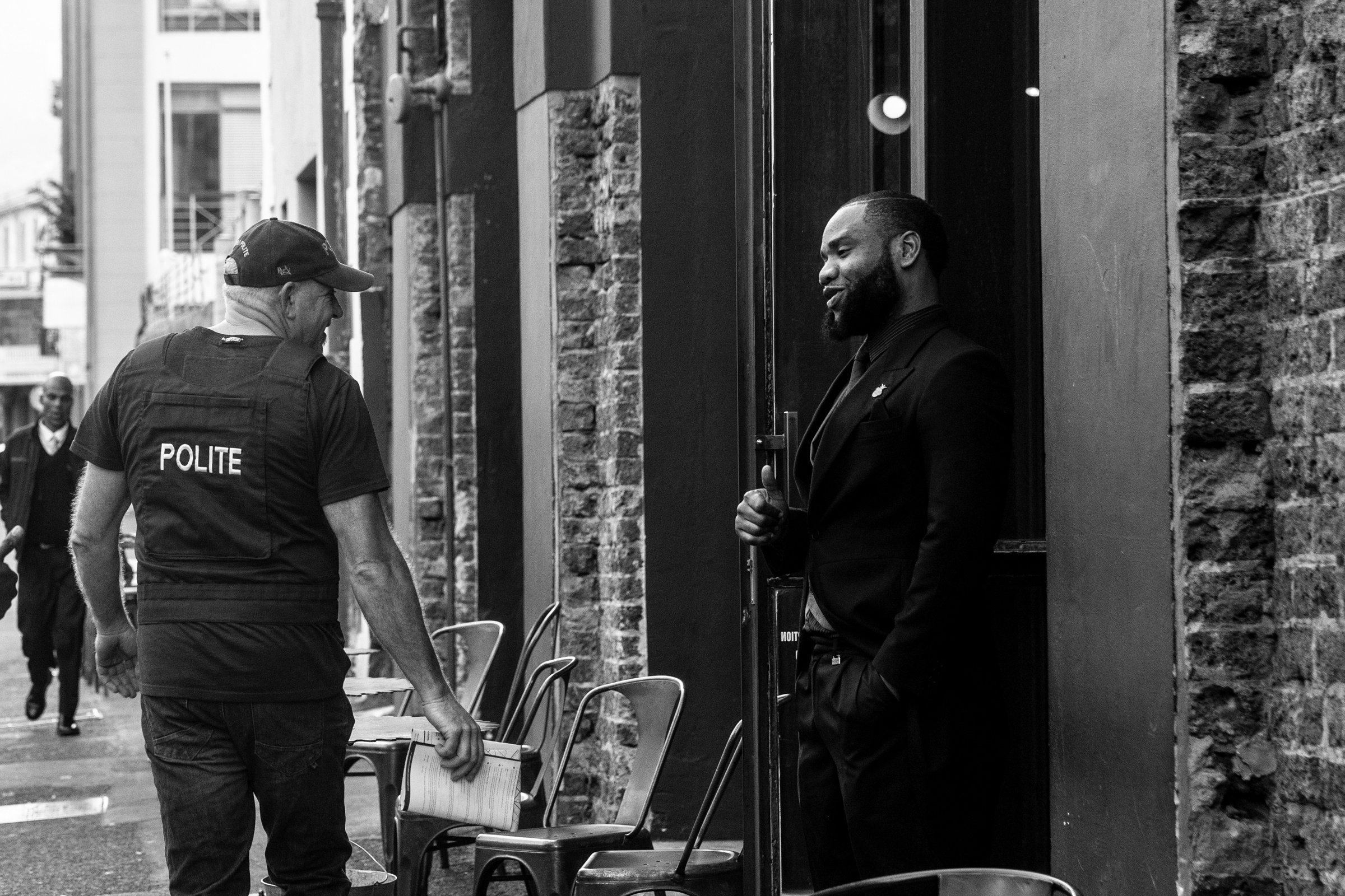 Monochrome process photograph from the 2019 rendition of Christian Nerf’s performance piece ‘Polite Force.’ On the left, a participant dressed in Polite Force riot gear is walking down a street in Cape Town. On the right, a employee of a business posted outside the entrance gives the participant a thumbs-up.
