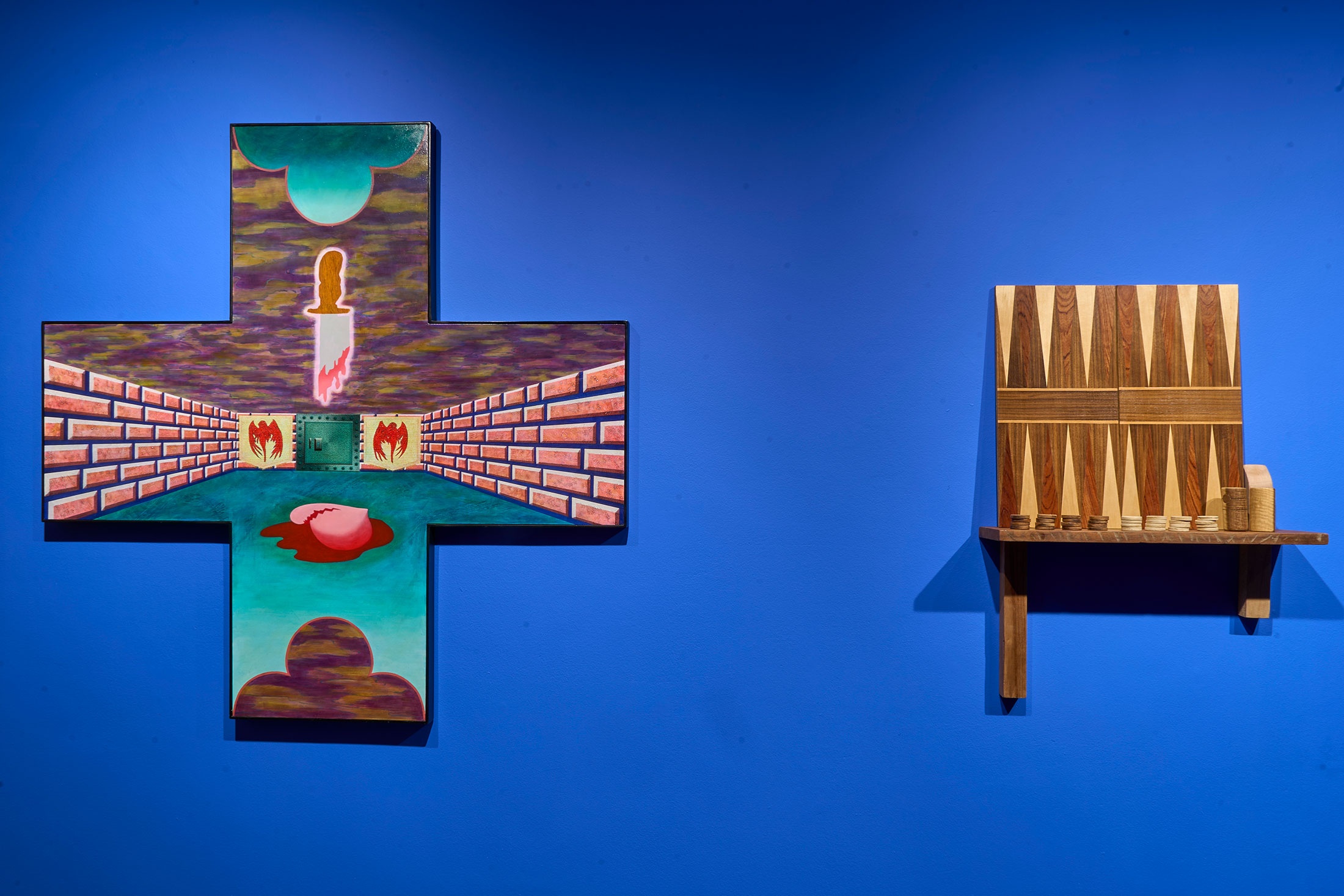 Installation photograph from the ‘Funhouse’ exhibition in A4’s Goods project space. On the left, Gitte Möller’s ‘Pushy Passion,’ oil and collage on a panel in the shape of an equal-armed cross. On the right, Rowan Smith’s ‘Untitled (Backgammon),’ a sculptural work made from carved wood that resembles a backgammon board and set pieces.
