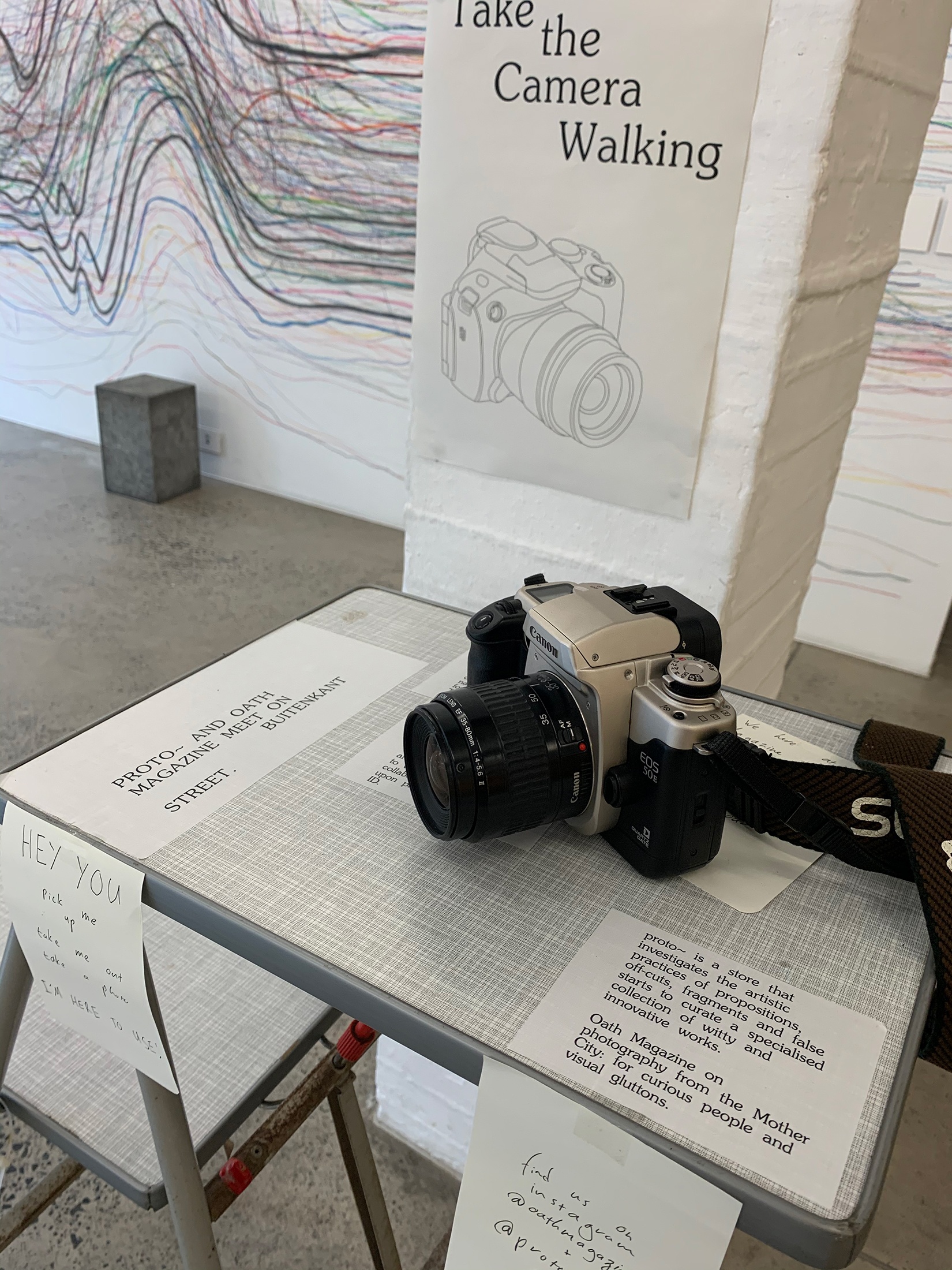 Event photograph from ‘Contact’, a collaboration between A4’s proto~ museum shop and Oath Magazine. A camera sits on a small table with explainer text. A poster on the pillar behind it reads “Take the Camera Walking”.
