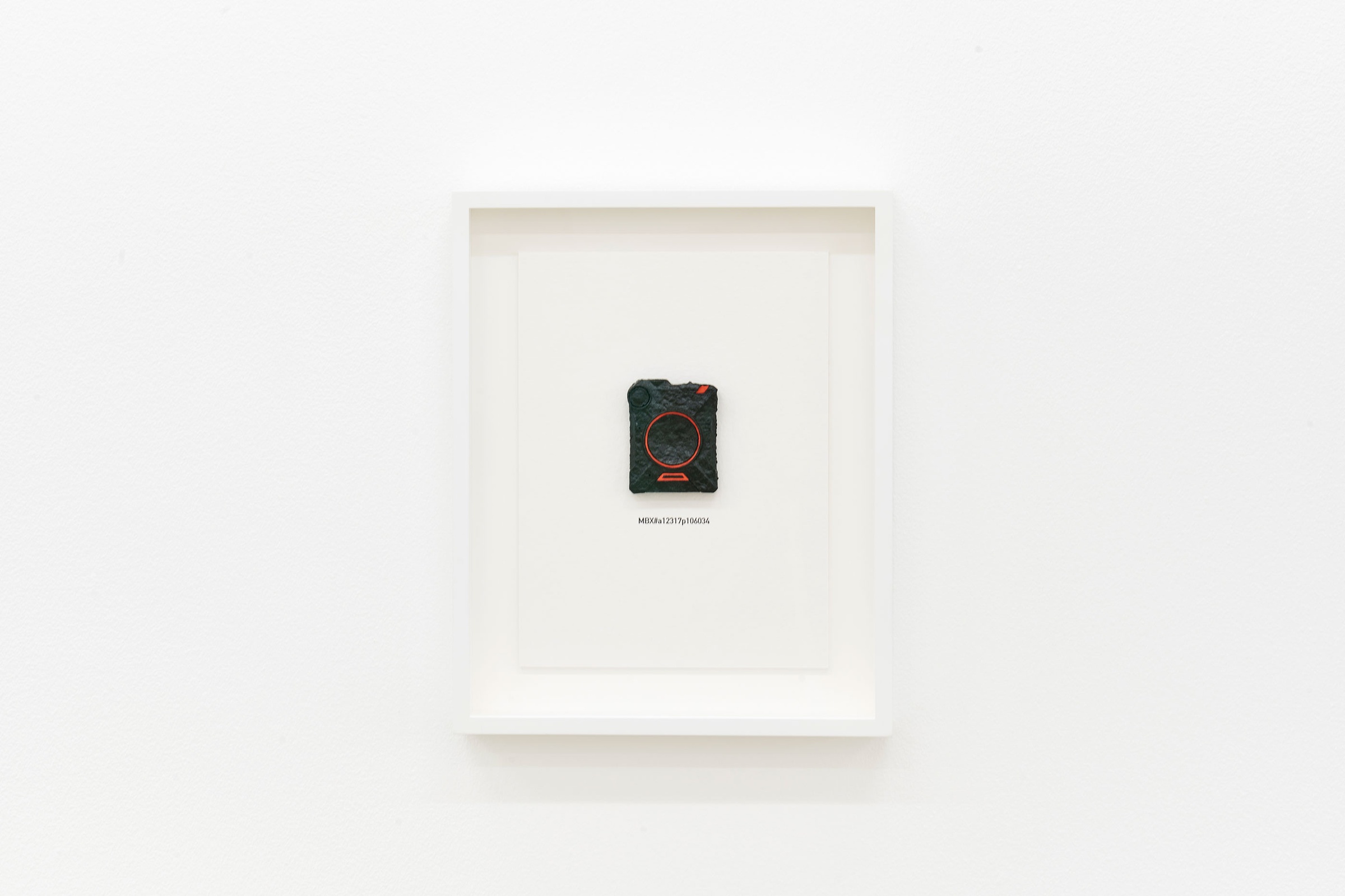 Installation photograph of the Common exhibition. Mark Bradford’s ‘Life Size’, a framed handmade reproduction of a policeman’s body camera is hung on a white wall.
