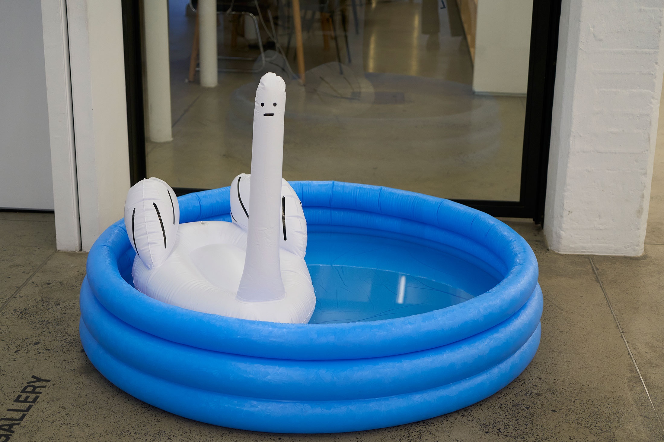 Event photograph from the launch of David Shirley’s ‘Inflatable swan thing’ in A4’s Proto~ museum shop. A white plastic inflatable swan-like creature floats in a blue inflatable children’s swimming pool.
