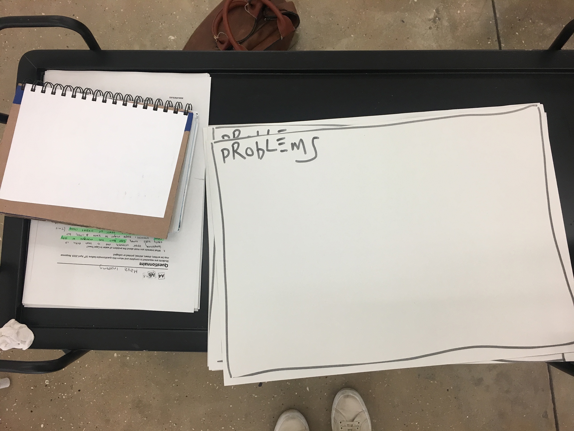 Photograph from the ‘Water Workshop with Edu Africa’ exchange in A4’s Gallery that depicts a black metal trolley. On the left, a stack of questionnaires and a paper notebook. On the right, a stack of papers with frames and the word ‘problems’ drawn in black felt marker.
