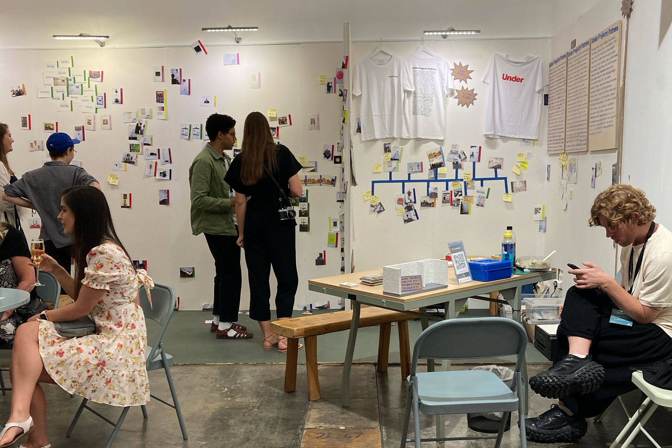 Event photograph from the Under projects booth at the 2023 Investec Cape Town Art Fair. White panelled walls are covered with printed snippets from the project archives, with team members and participants standing in discussion.
