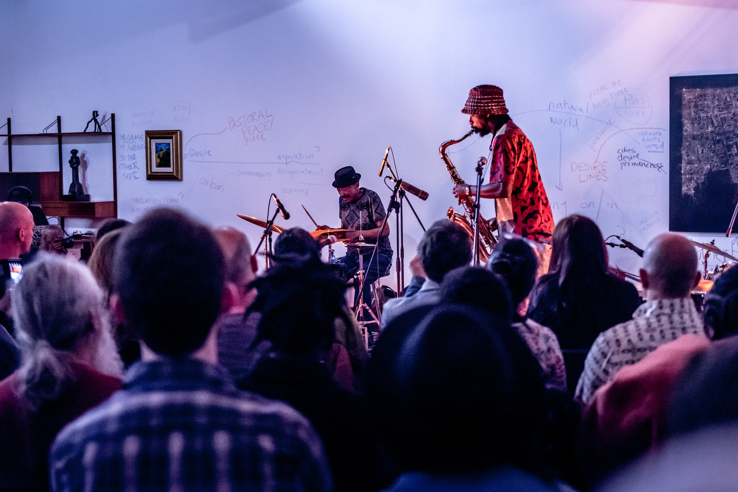 Event photograph from a performance by Shabaka and the Ancestors, with special guest Bra Louis Moholo-Moholo, on A4’s top floor. At the back, the musicians are performing in front of a white wall that features text and drawings, with Gerard Marx’s mixed media work ‘Prayer Carpet: Aerial’ mounted on the wall on the right. At the front, attendees are seated.
