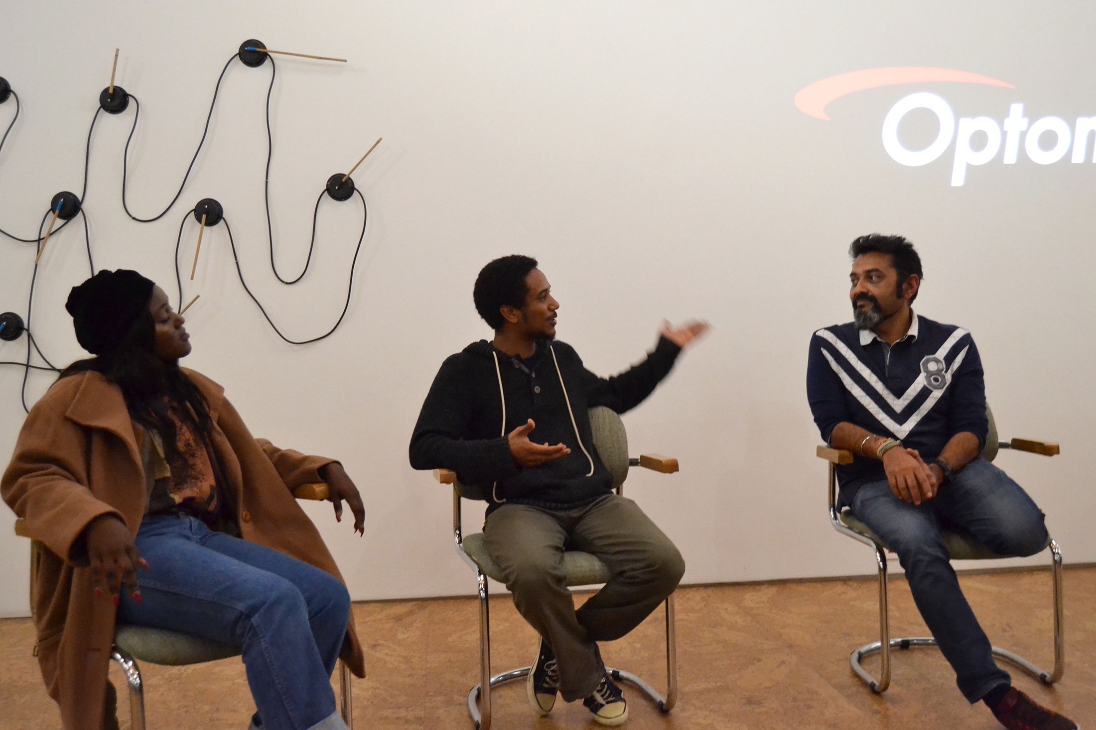 Event photograph from the 'AFDA and A4 Exchange: Experimental Film and Video Art’ on A4’s top floor. At the front, moderators heeten bhagat and Tabogo Nong sit with a participant. At the back, Josh Ginsburg’s kinetic wall sculpture ‘Science fiction (Gauges)’ is mounted on a white wall.
