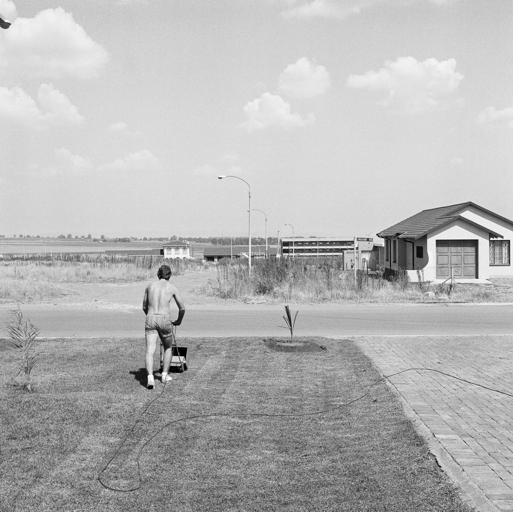 David Goldblatt's monochrome photograph 'Saturday afternoon in Sunward Park, Boksburg' shows a shirtless individual mowing a lawn.
