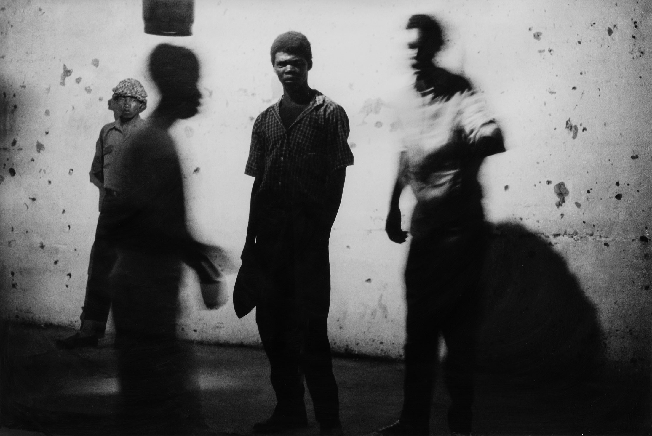 Santu Mofokeng’s monochrome photograph ‘Concert at Sewefontein, Bloemhof’ depicts four individuals standing in front of a wall.

