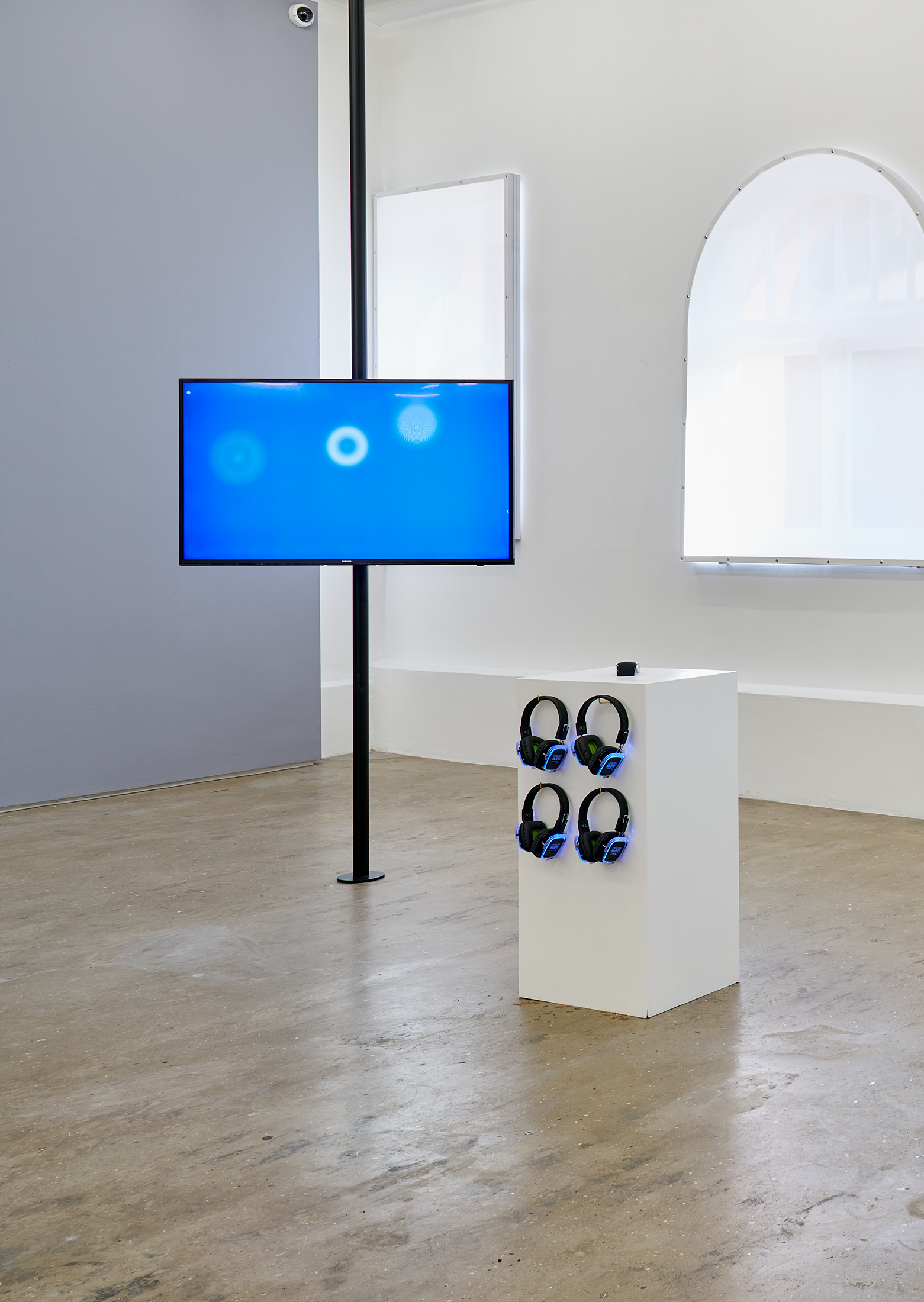 Installation photograph from the ‘Sounding the Void, Imaging the Orchestra V.1’ exhibition in A4’s Gallery. Jenna Sutela and Ville Haimala's web-based work consists of a screen mounted on a vertical metal beam on the left, and plinth with four sets of headphones on the right.
