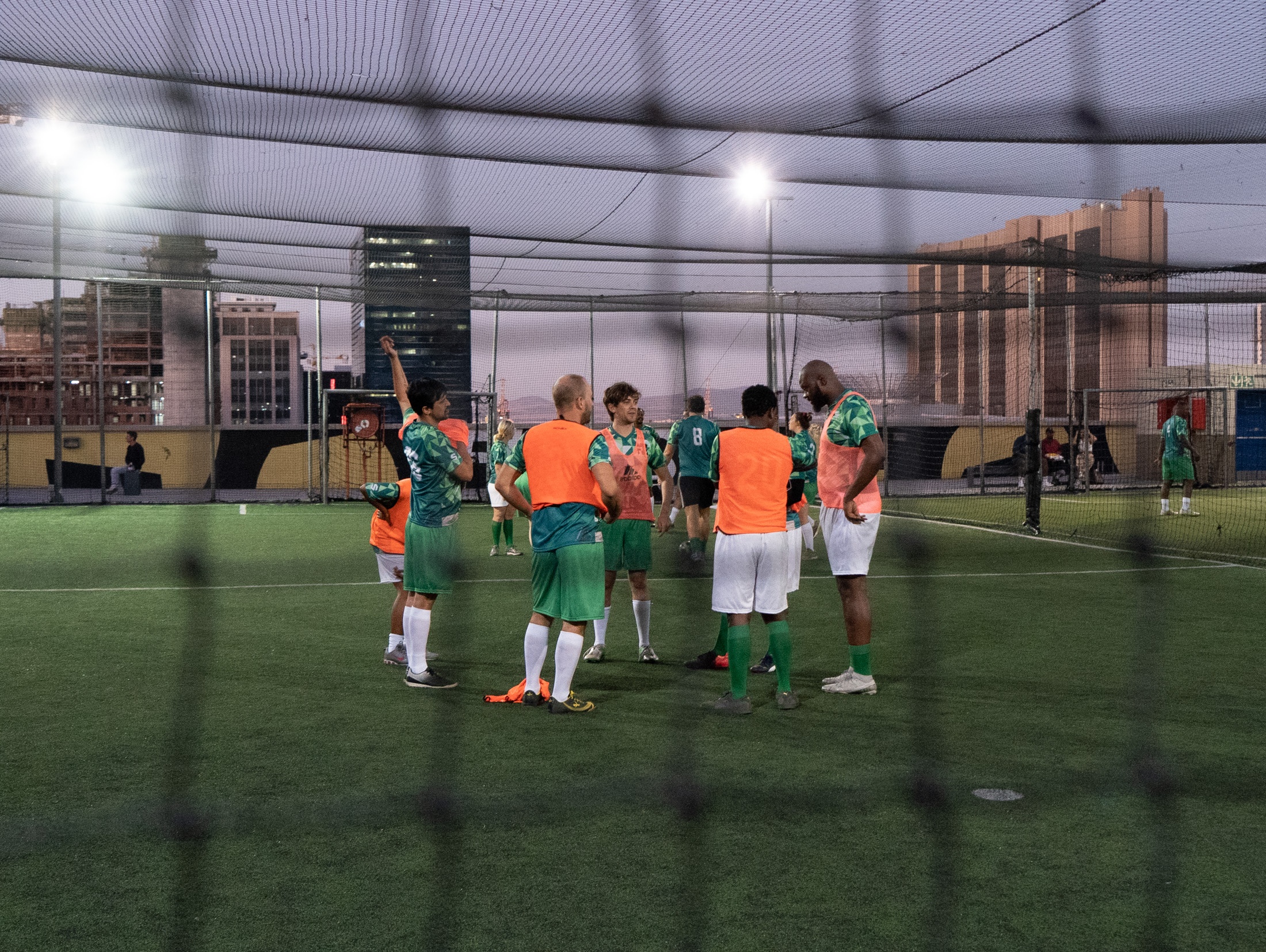 Event photograph from the 2023 edition of Exhibition Match depicts participants talking on a soccer field on a lit rooftop field in the evening.
