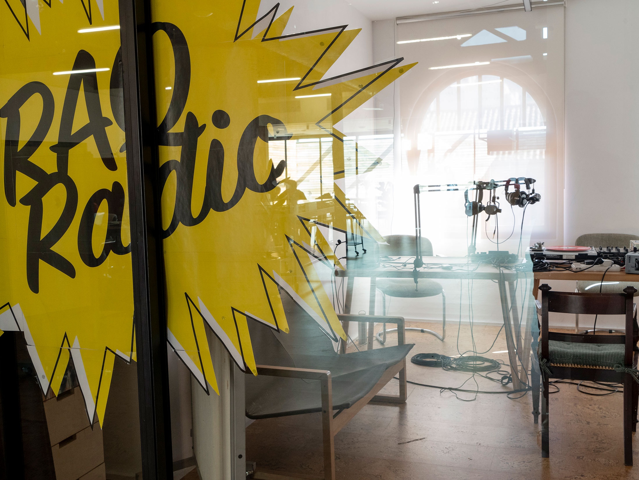 Process photograph from the RAQ Radio residency in A4's second-floor studio. At the front, the black and yellow RAQ Radio logo is pasted onto the transparent sliding doors. At the back is a long wooden table equipped with earphones, microphones, an audio mixer and turntable.
