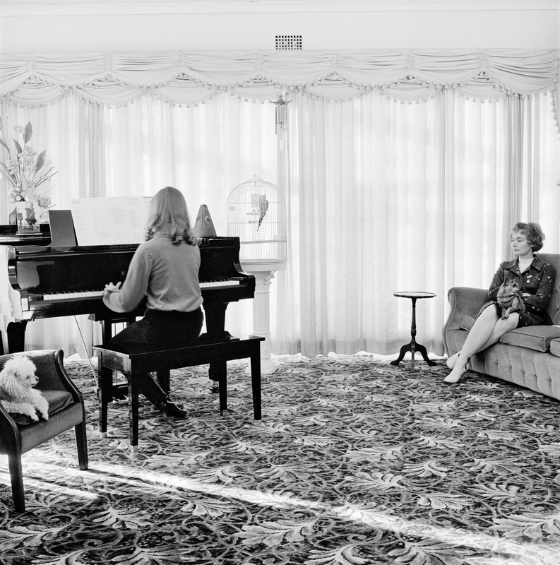 David Goldblatt's monochrome photograph 'A girl and her mother at home, Boksburg. 22 June 1980' shows a young individual playing a piano on the left, and an older individual sitting on a couch on the right.
