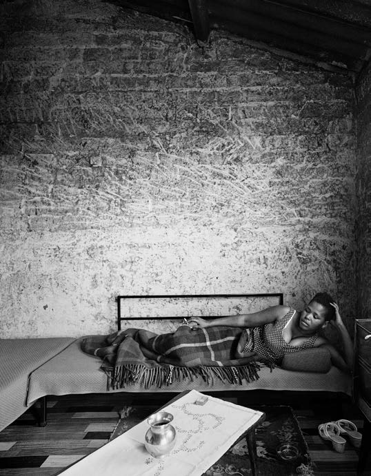 David Goldblatt's monochrome photograph 'Margaret Mcingana who later became famous as the singer Margaret Singana, Zola, Soweto' shows an individual lying on a bed at the base of a brick wall, smoking a cigarette.
