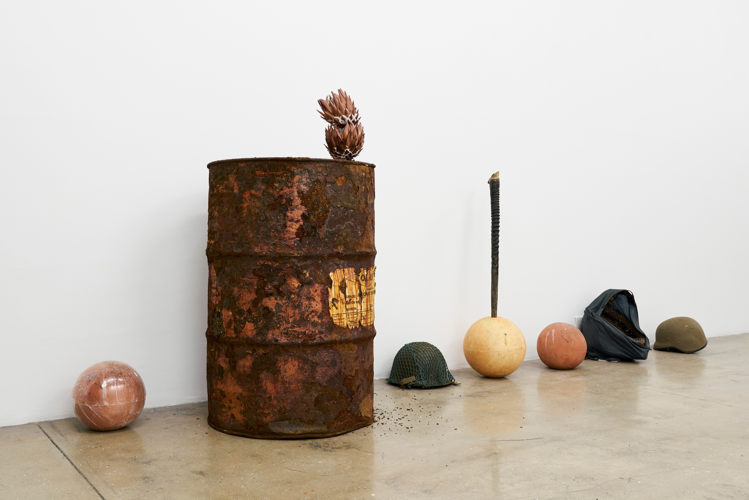 Artwork photograph from the ‘without a clear discernible image’ exhibition in A4’s Gallery that depicts Kevin Beasley’s untitled installation. At the back, a series of object are arrange along the base of the gallery wall, including an oil drum with protea flowers protruding from it’s opening, a ceramic yard globe with an antelope horn protruding from it and military helmet sitting within a handbag.
