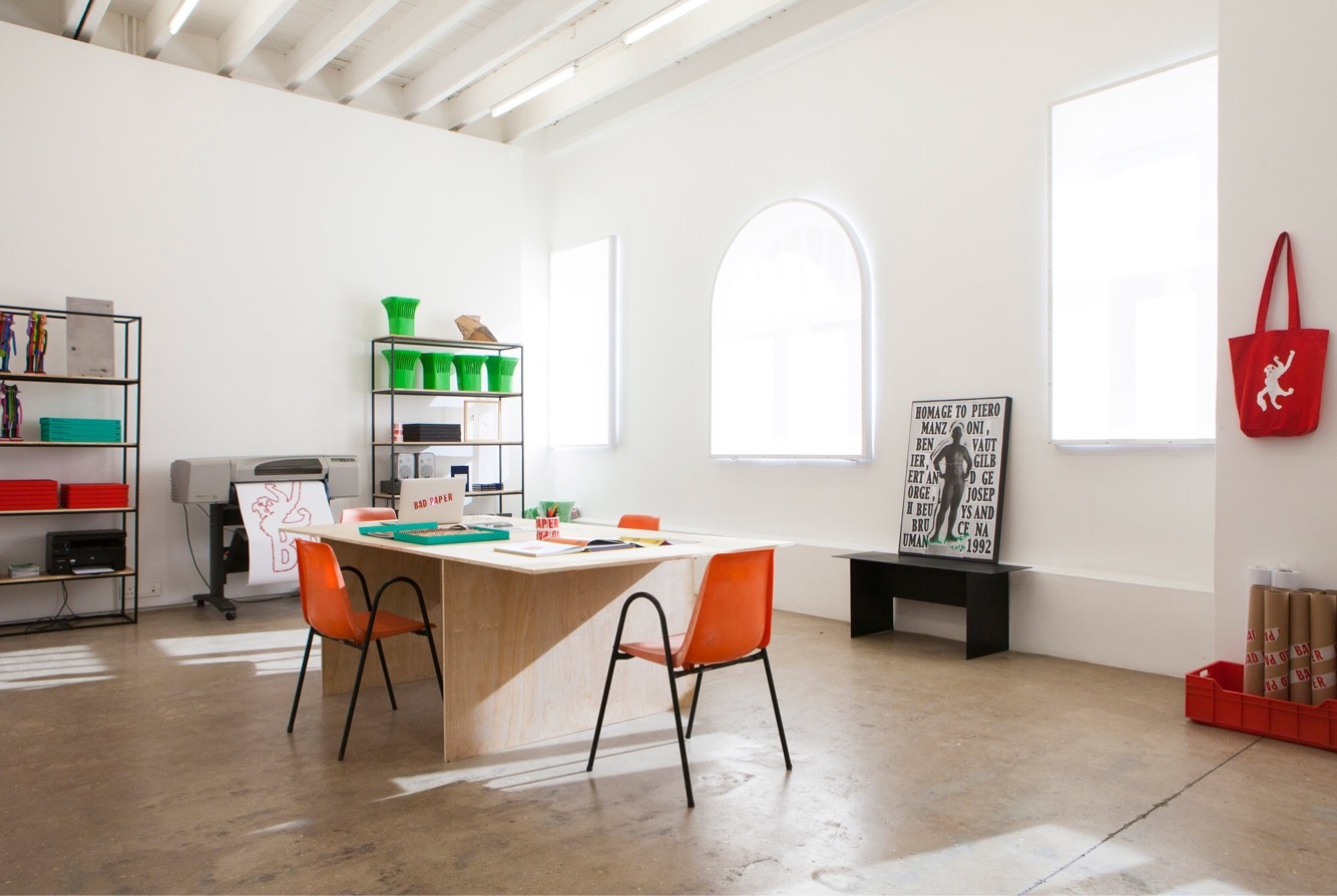 Installation photograph from the Bad Paper residency on A4’s top floor. In the middle, a table with seating that features books and a laptop. At the back, two shelving units and a printer.
