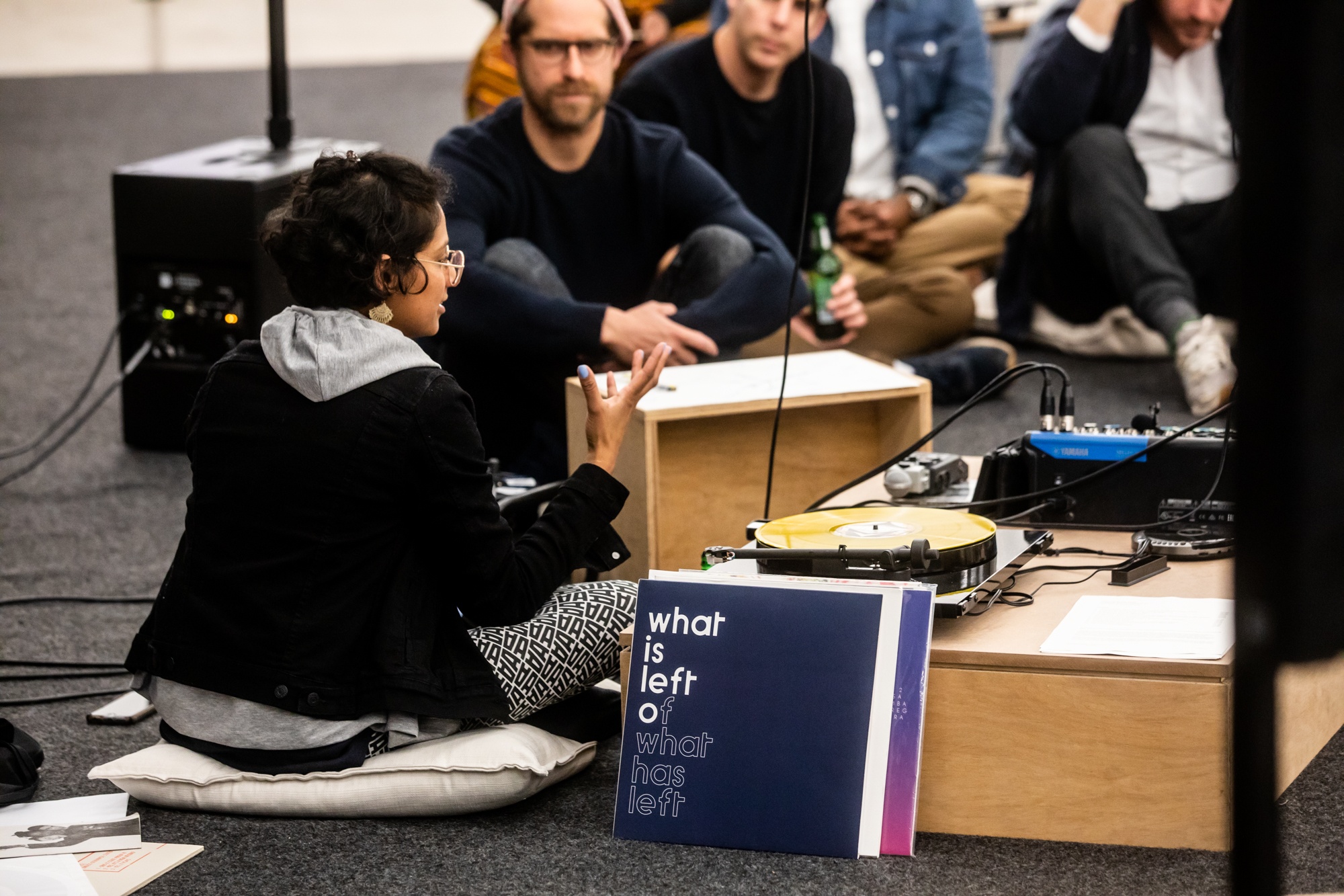 Event photograph from ‘How we listen determines what we hear,’ a conversation between Bhavisha Panchia and Josh Ginsburg, in A4’s Gallery. On the left, Panchia and Ginsburg are seated on the gallery floor. On the right, audio equipment installed for the ‘Sounding the Void, Imaging the Orchestra V.1’ exhibition.
