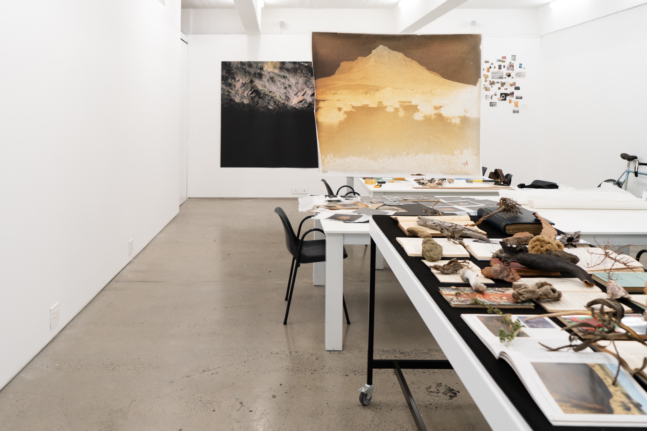 A process photograph from Tommaso Fiscaletti and Hemel Liggaam's residency at A4 Arts Foundation, showing the unpacking of their archive. On the right, a row of white tables hold books, printouts and objects from the archive. In the middle, a photographic print is suspended from the ceiling. At the back, photographic prints are mounted on a white wall.
