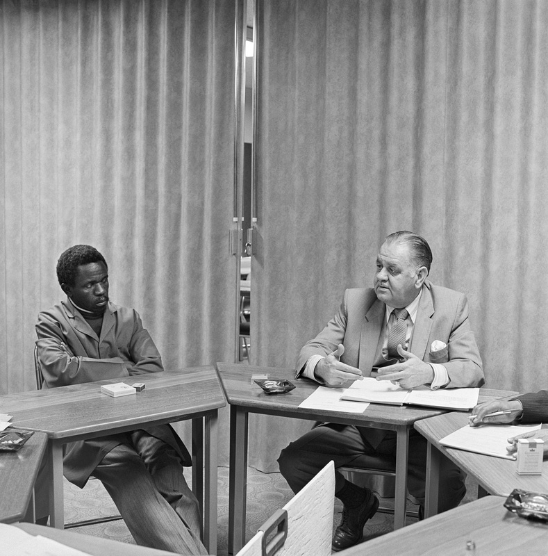 David Goldblatt's black-and-white photograph 'At a meeting of the Worker-Management Liaison Committee of the Colgate-Palmolive company, Boksburg' shows two men conversing behind tables.
