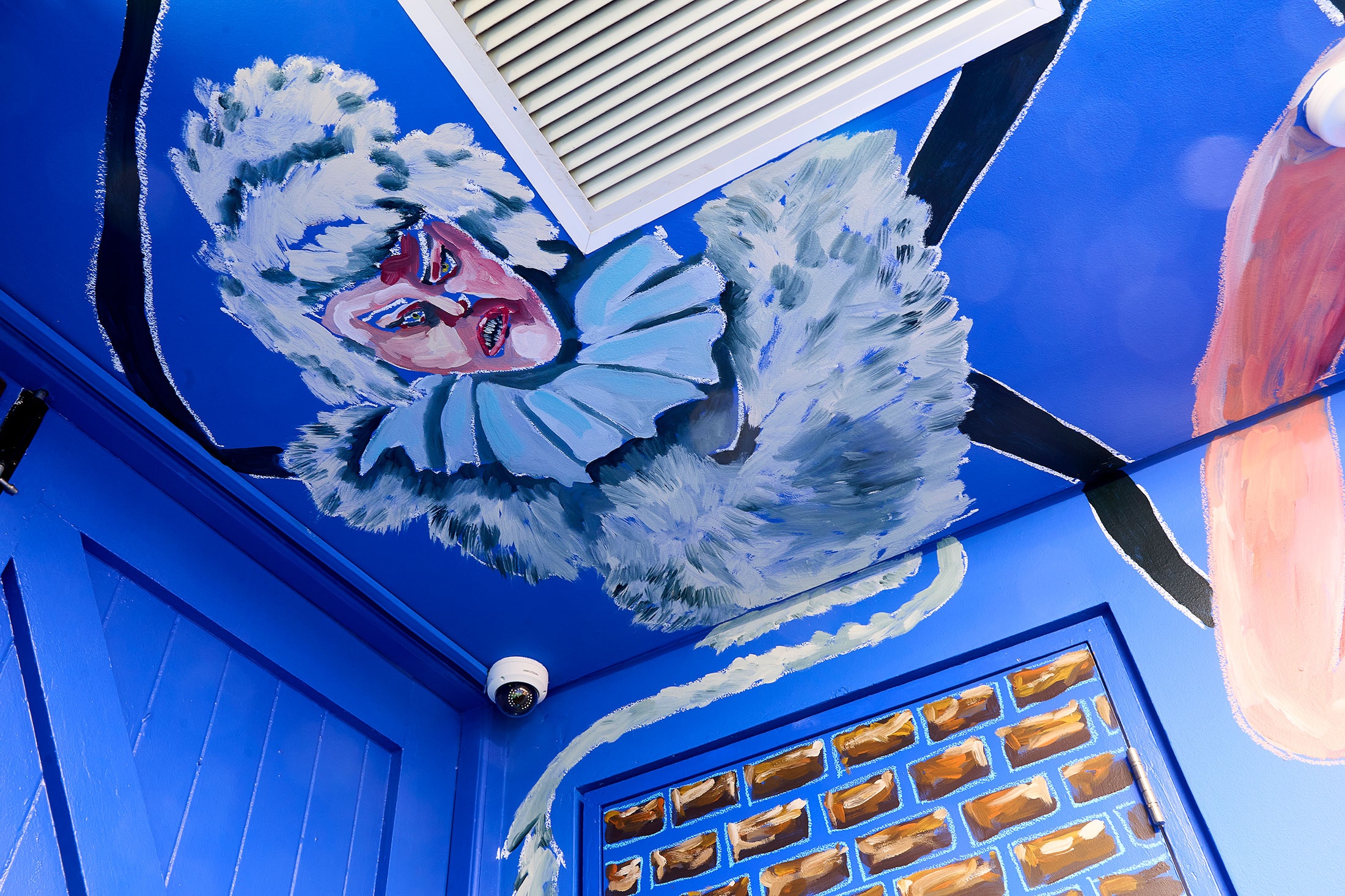 Installation photograph of Dominique Cheminais’ residency in A4’s Goods project space. A detail of a painted mural on a blue wall shows a flailing humanoid figure adjacent to the air vent.
