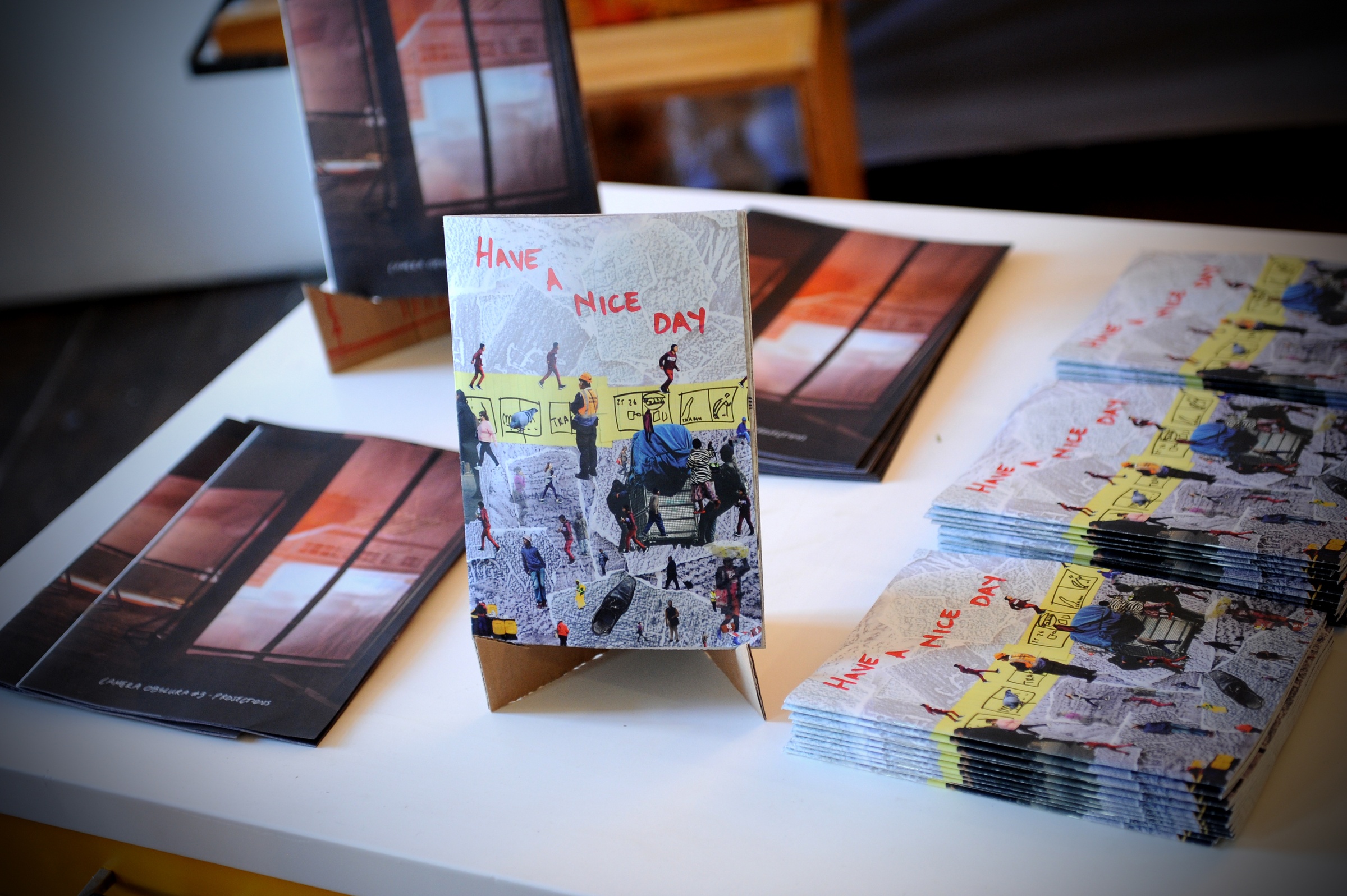 Event photograph from the offsite ‘Office Politics’ exchange. Multiple copies of the ‘Have a nice Day’ publication by Andrew Juries and Lauren Theunissen and the ‘Camera Obscura #3’ publication by Tebogo George Mahashe are arranged on a white table.
