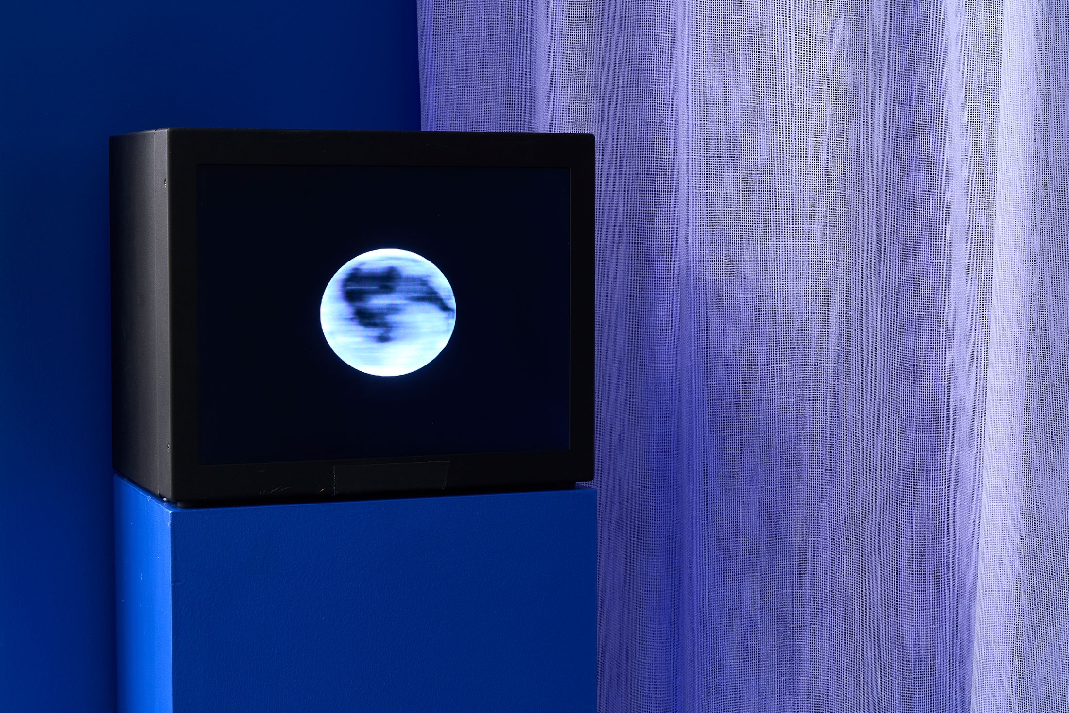 Installation photograph from the ‘Lunar Maria Chorus’ exhibition in A4’s Goods project space. On the left, a black box on a blue plinth displays a video of the moon rotating. On the right, a curtain of white gauze.
