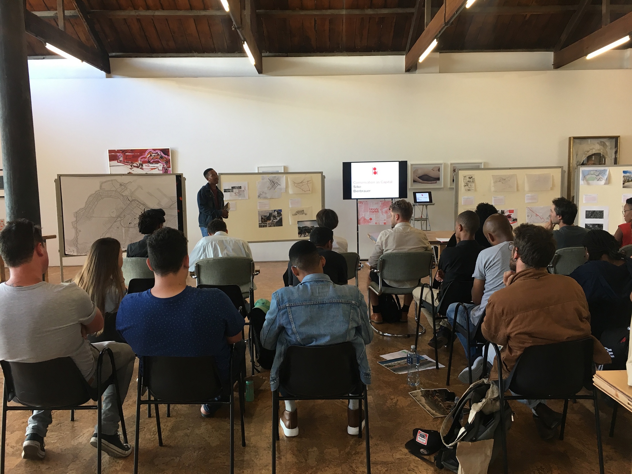 Process photograph from the ‘Unit 11 - Bloody Agency’ exchange with students from the University of Johannesburg’s Graduate School of Architecture on A4’s top floor. At the back, a series of pinboards and a screen accompany a presentation by an individual. At the front, audience members are seated in rows.
