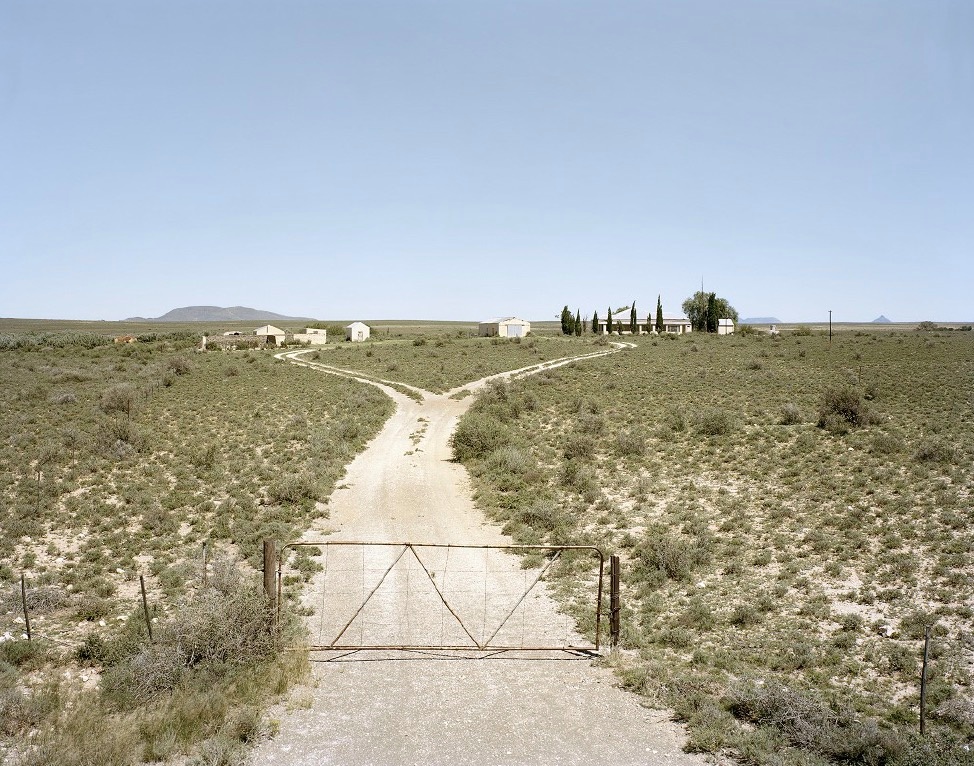 David Goldblatt's photograph 'Deserted farm. Holgatsfontein in the Leeukopspan area, between Britstown and Vosburg, Northern Cape' shows a single dirt road closed off by a gate that splits into two diverging roads at the back.
