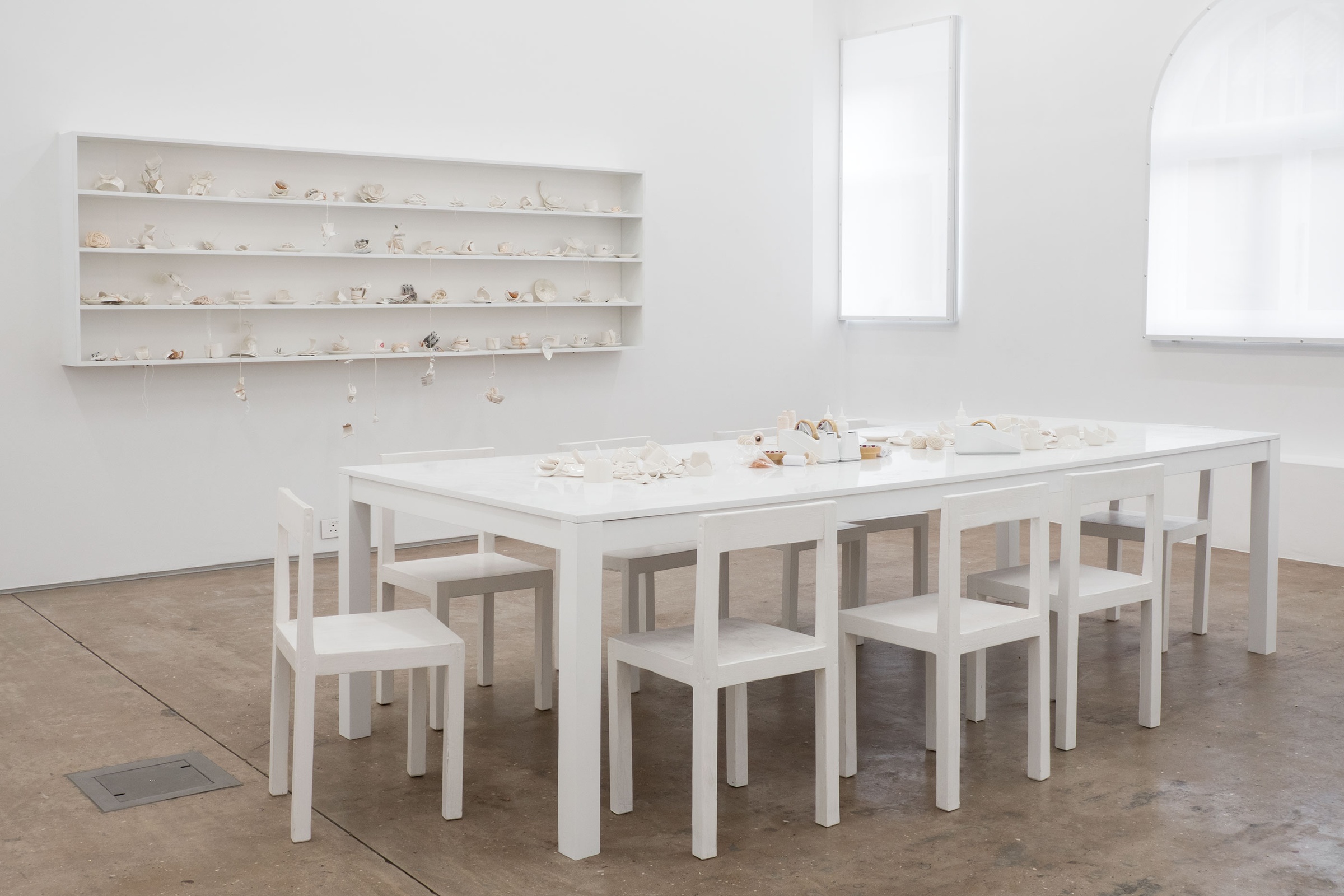 An installation photograph of Yoko Ono's installation 'MEND PIECE, Andrea Rosen Gallery, New York City version'. At the front, a white table surrounded by chairs holds ceramic fragments, glue, tape, scissors and twine. At the back, a white wall-mounted shelf holds pieces constructed by participants.
