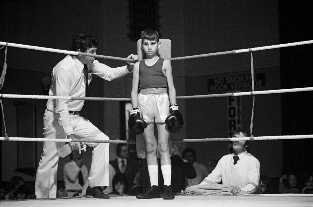 David Goldblatt's black-and-white photograph 'Before the fight: amateur boxing at the Town Hall, Boksburg. 1980' shows a young boy positioned in the centre of the frame, standing in a boxing ring. There is a man standing on either side of him.
