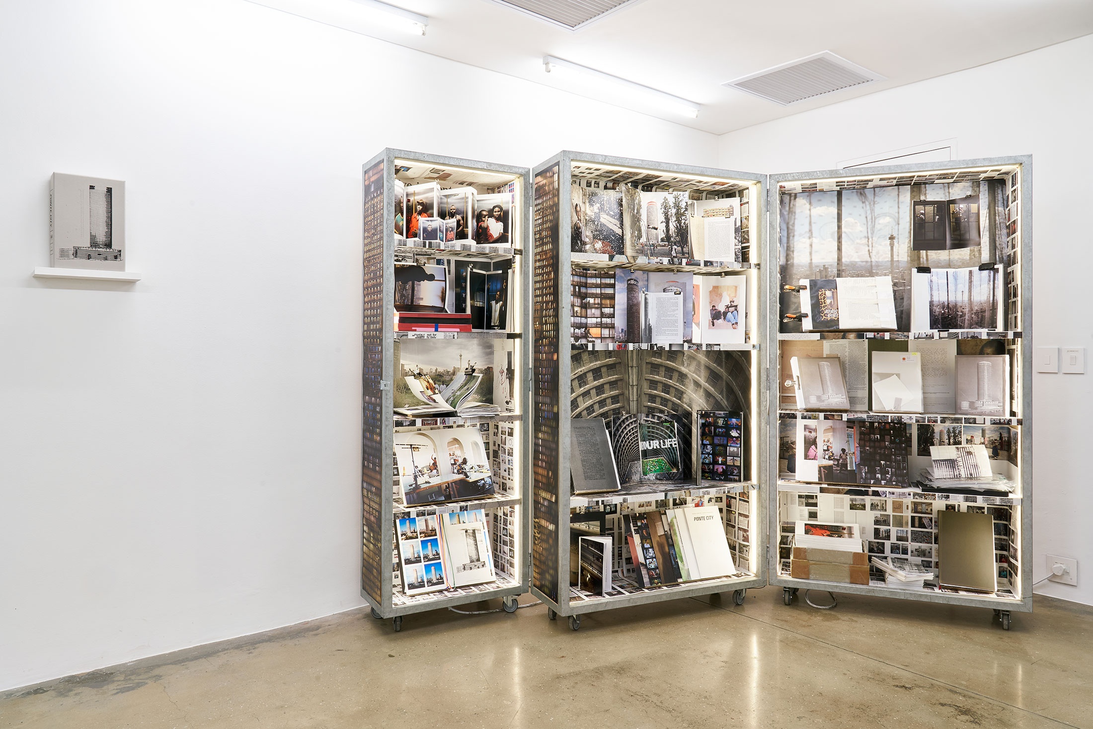 Installation photograph from the Photo Book! Photo-Book! Photobook! exhibition in A4’s Gallery. On the right, Mikhael Subotzky and Patrick Waterhouse’s cabinet installation of the 'Ponte City' archive. On the left, Mikhael Subotzky and Patrick Waterhouse’s book ‘Ponte City’ sits on a wall-mounted shelf.
