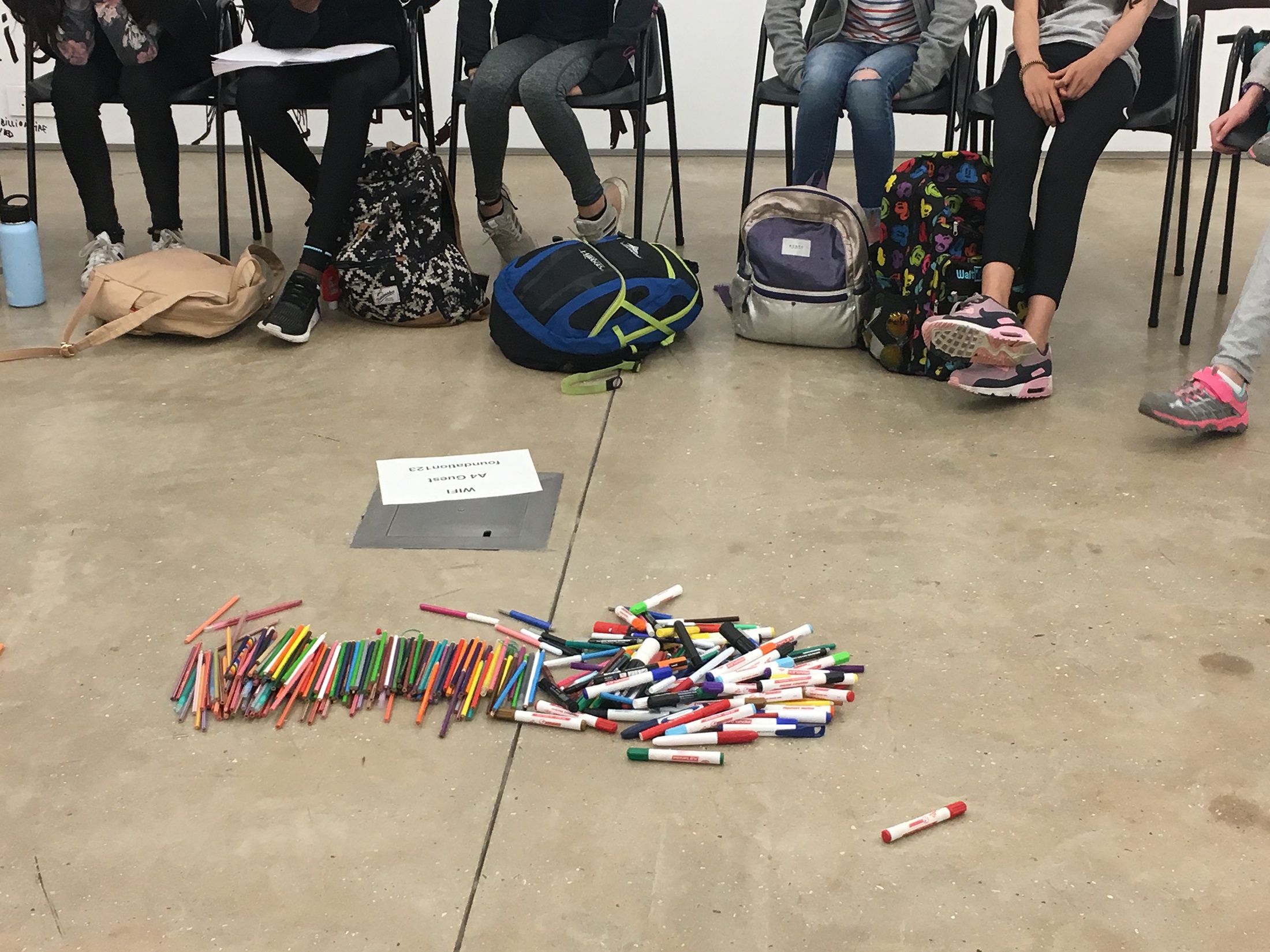 Photograph from the ‘Water Workshop with Edu Africa’ exchange in A4’s Gallery. At the front, a pile of colour pencils and markers are on the floor. At the back, young students from the Avenues World School are seated on chairs.
