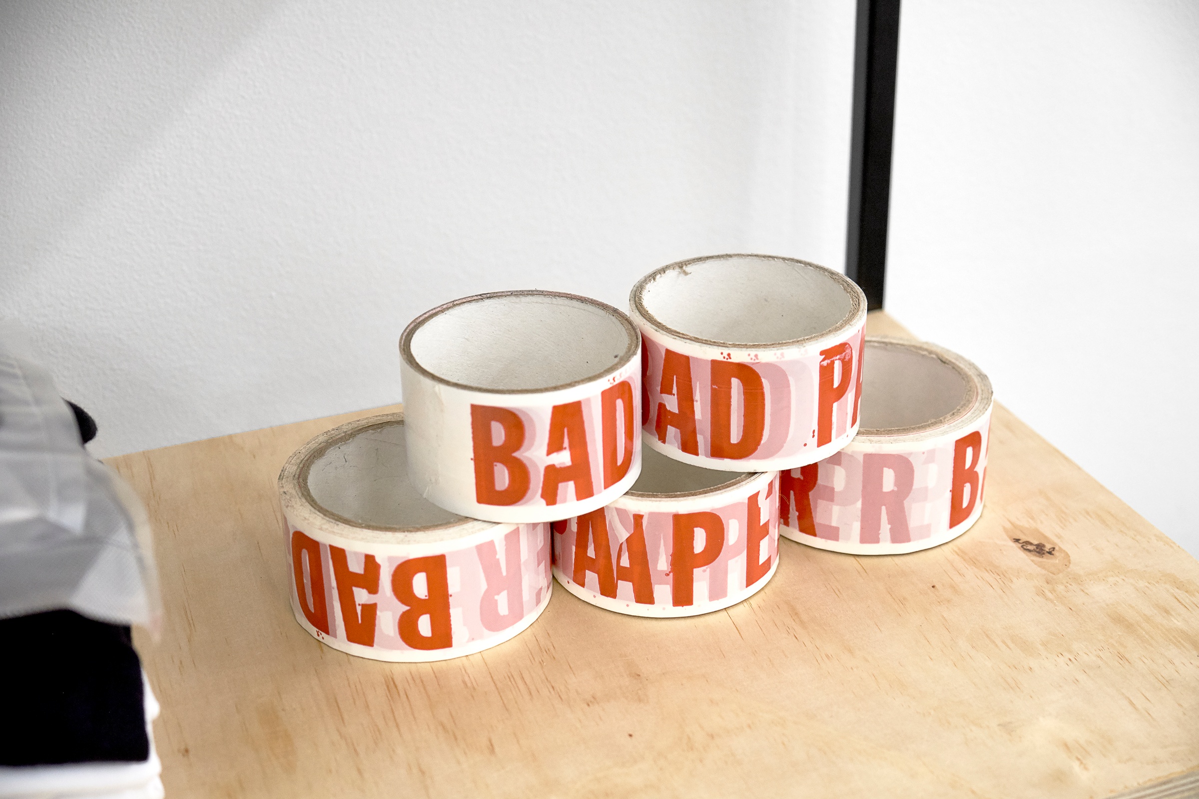 Installation photograph from the Bad Paper residency on A4’s top floor shows rolls of tape on a wooden shelf. Th semi-transparent white tape repeats the phrase ‘BAD PAPER’ in bold red font.
