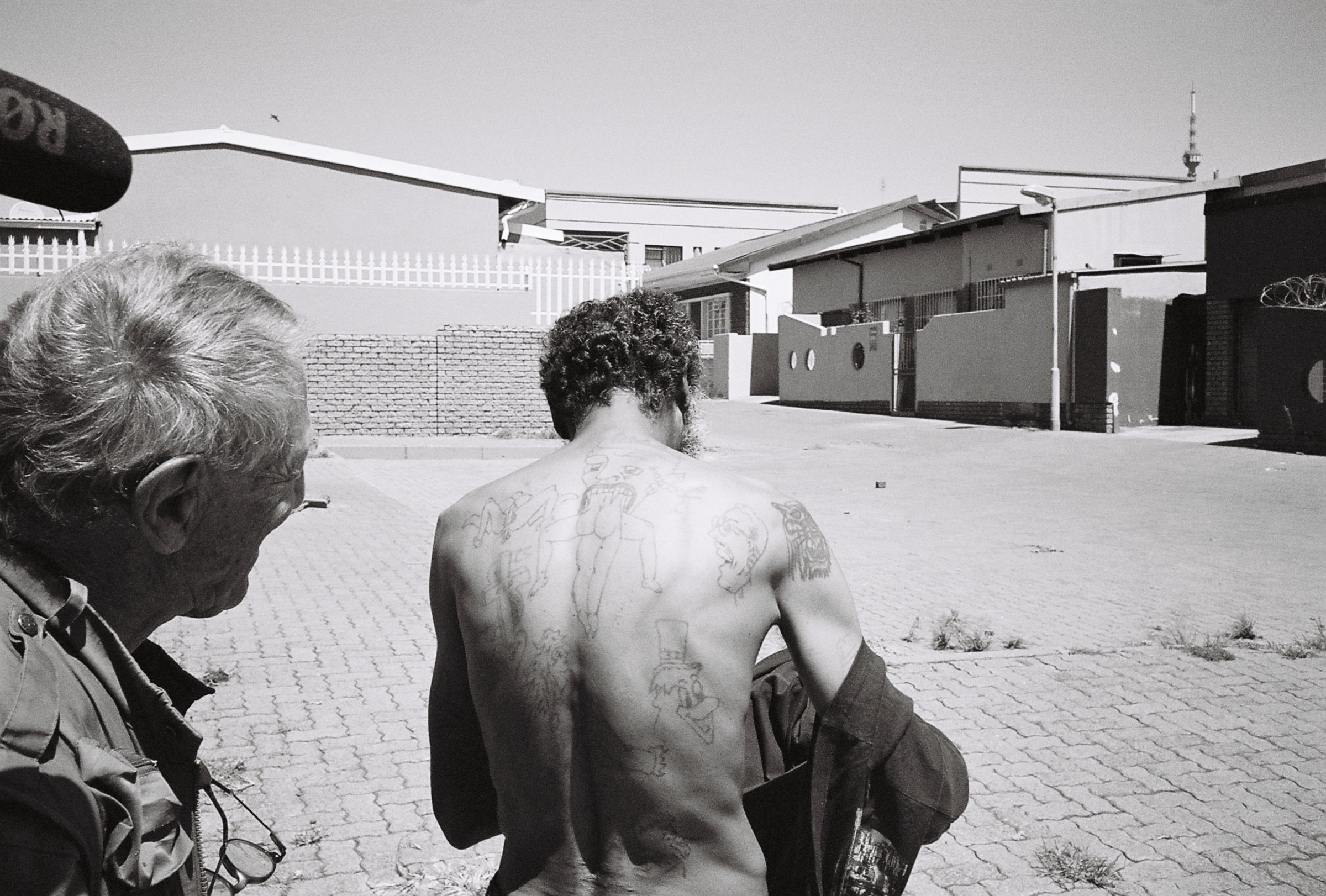 A monochrome process photograph taken in Fietas, Johannesburg during the making of the film ‘Goldblatt: A Documentary’ by director Daniel Zimbler. On the left, the side of David Goldblatt’s face. In the middle, a man with his back exposed, covered in tattoos. In the back, various buildings.
