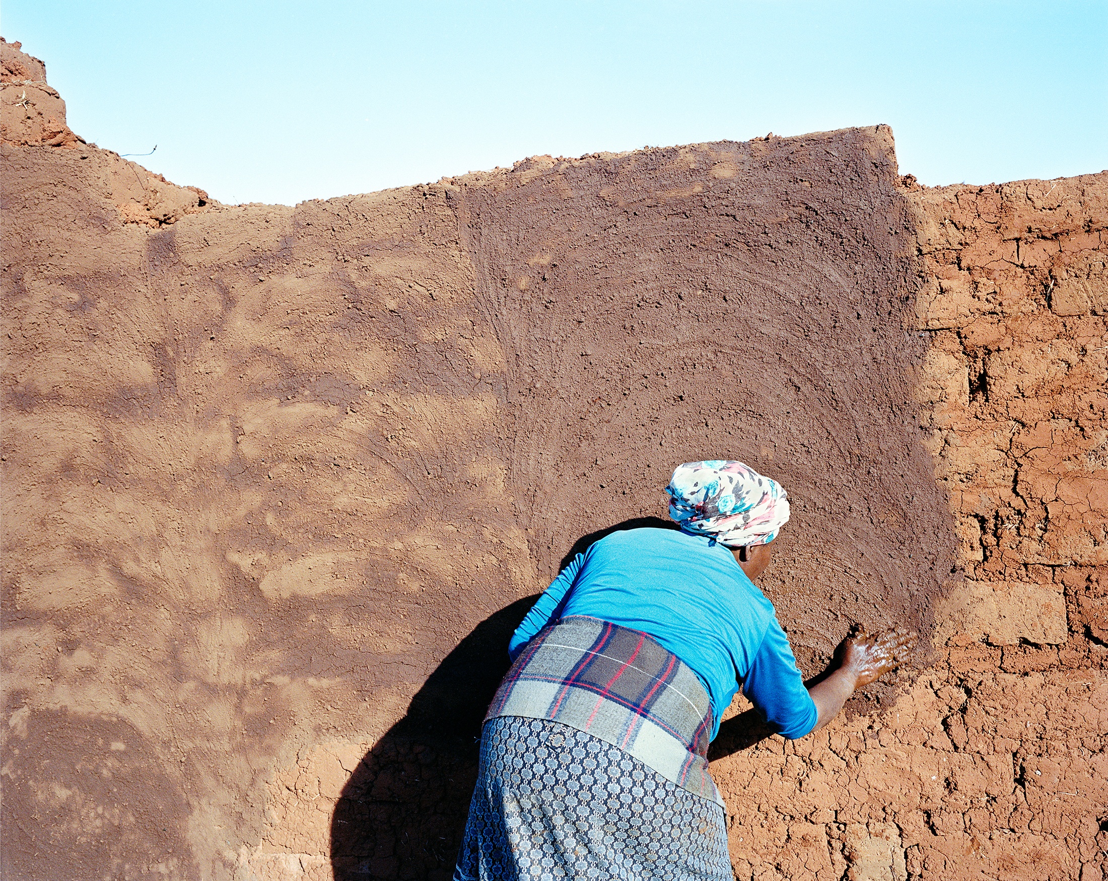 Lindokuhle Sobekwa's photograph 'UmamBhele uyatyabeka' shows an indvidual rubbing a brown substance onto a brick wall.
